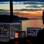 man looking at computer monitors reviewing stock trends with sunset in the background