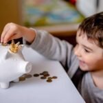 young child with smile on face while putting coin into piggy bank for savings